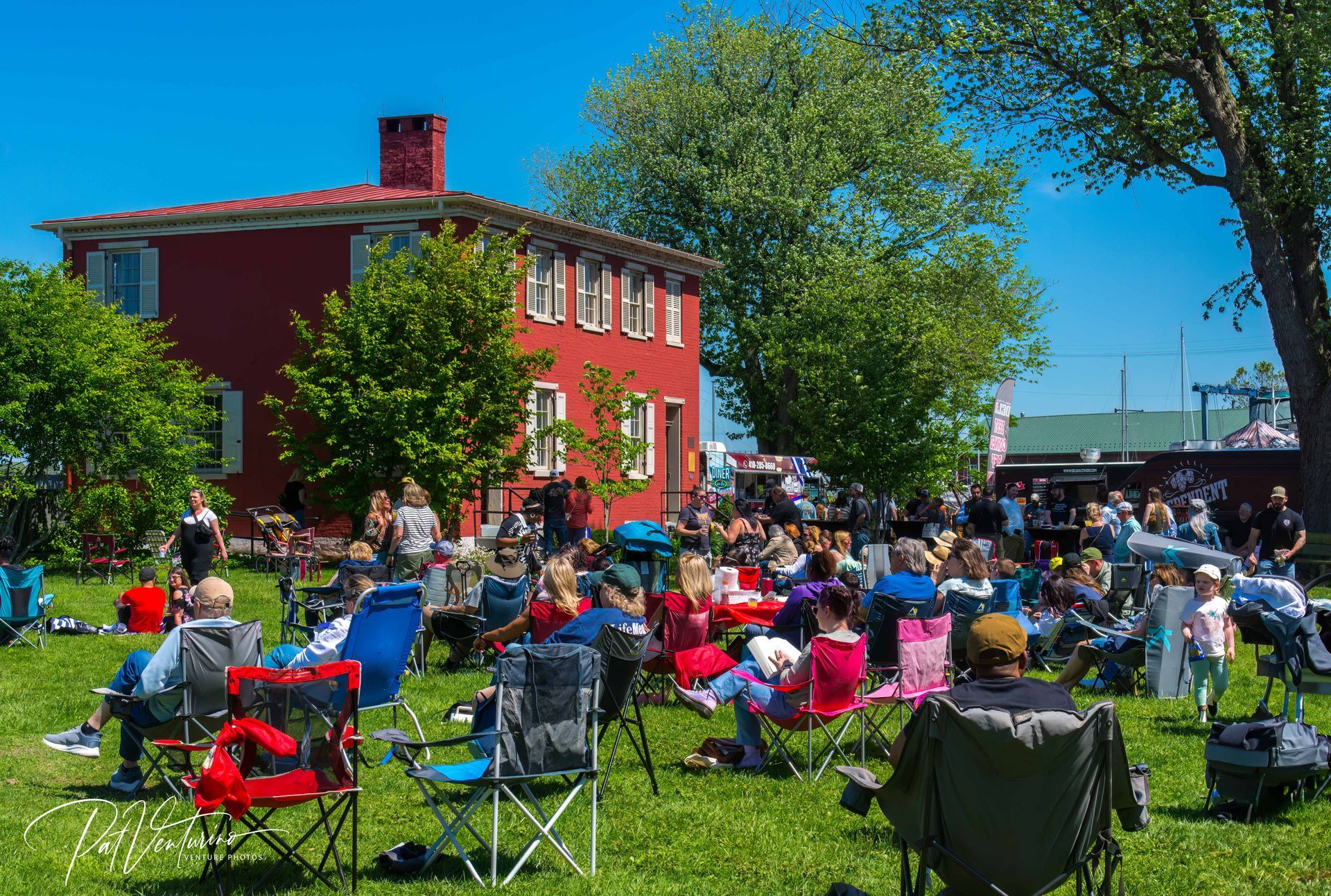 People sitting in chairs on grass, facing a red building with trees, likely at an outdoor event on a sunny day.