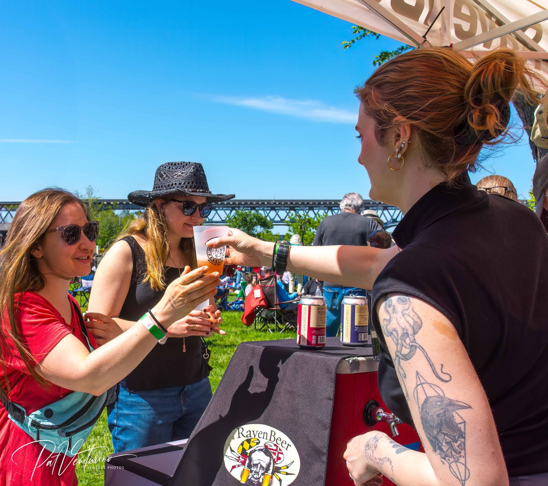 People at an outdoor beer festival. A woman serves beer. Others wear sunglasses, holding glasses.