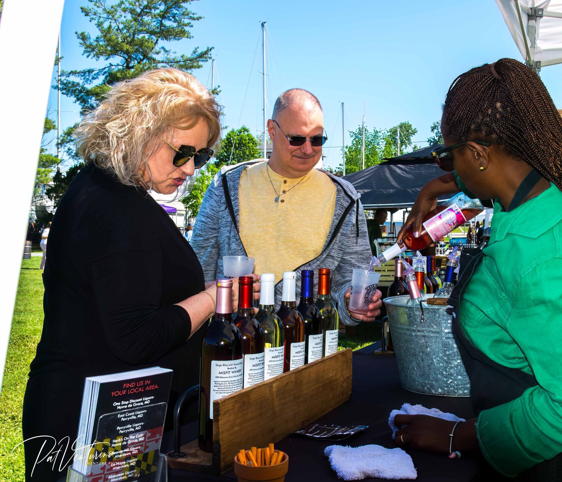 People at a wine tasting event. Woman pouring wine, others sampling; bottles of wine on a table, outdoors on a sunny day.