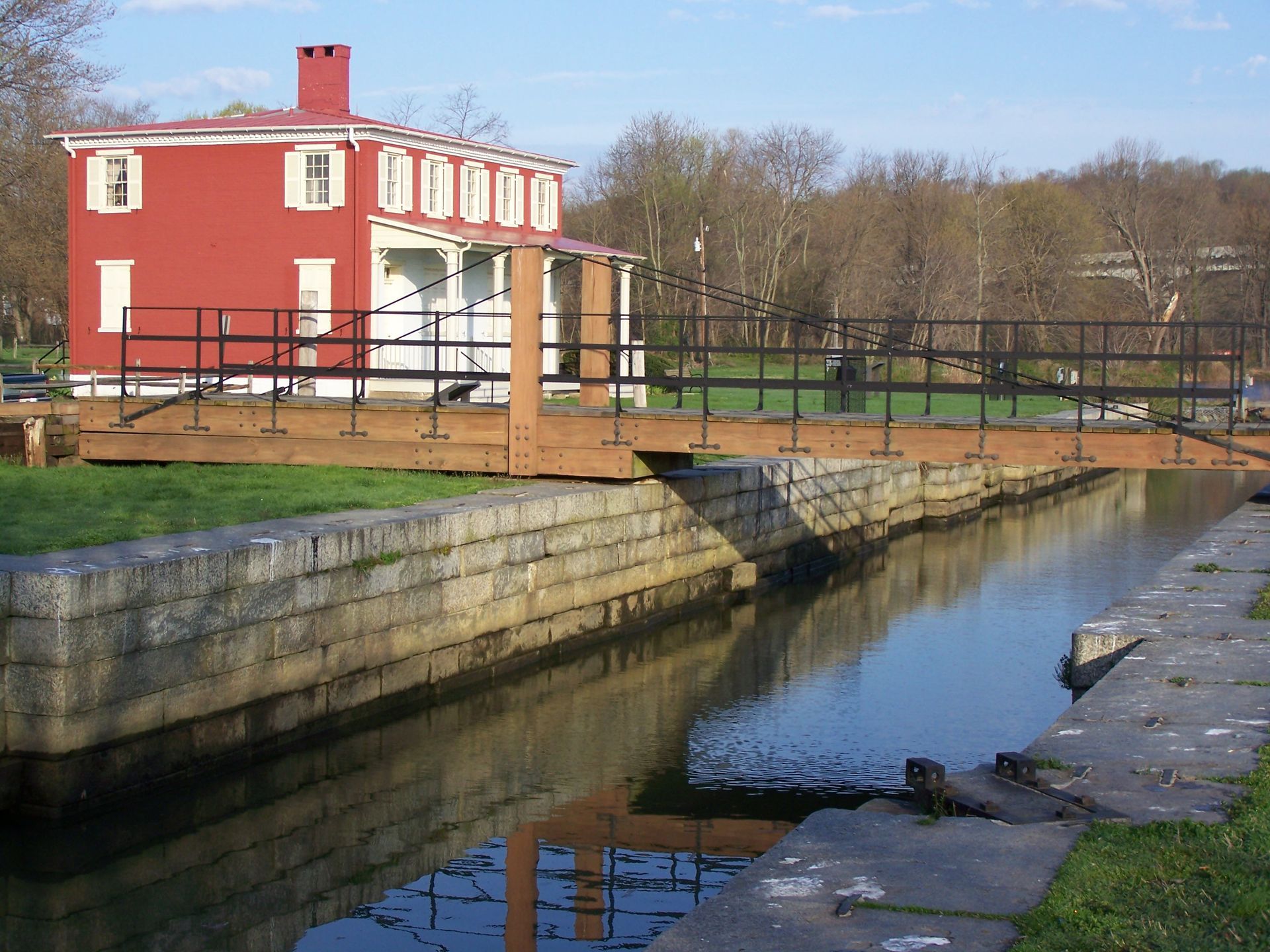Red building next to a canal with a bridge. Stone walls, grassy area, trees in the background.