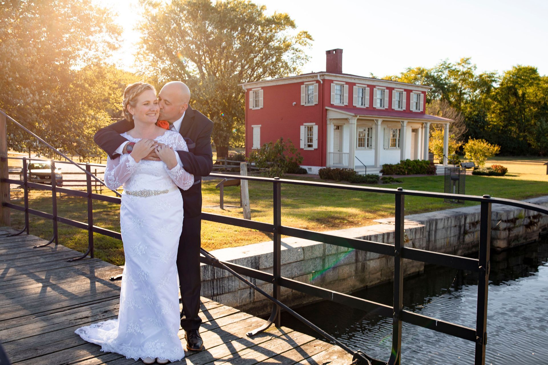 Bride and groom embrace on a bridge, red house in the background, warm sunlight.