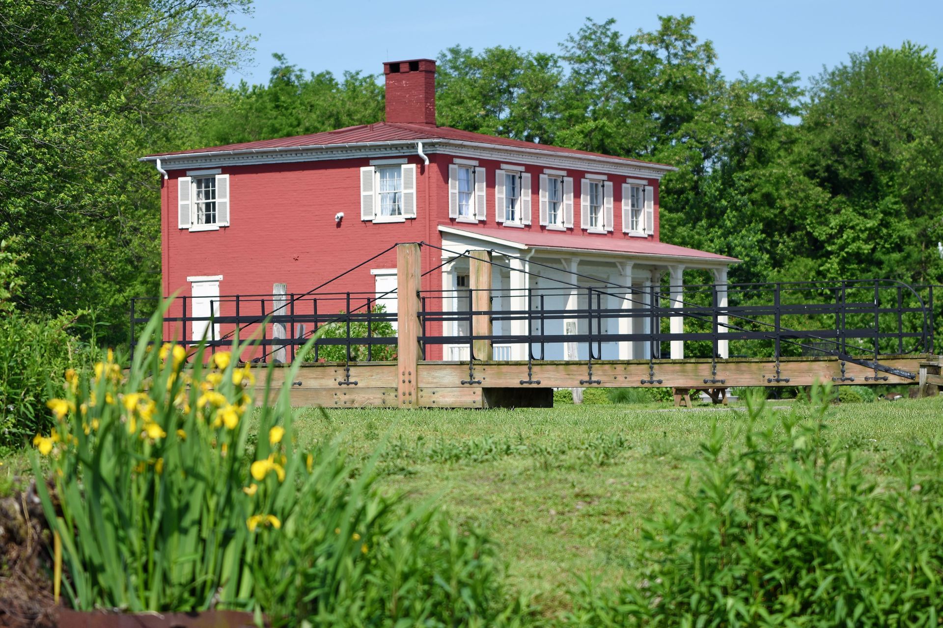 Red house with white trim and shutters, front porch, on a grassy area. Yellow flowers in foreground.