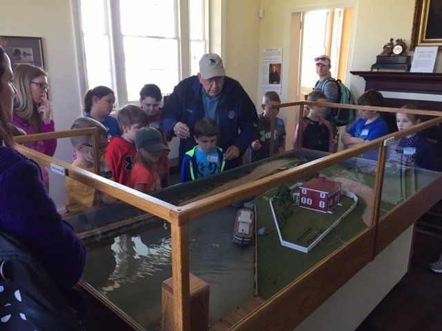 Children and an older man look at a model river landscape in a museum, focused on a boat.