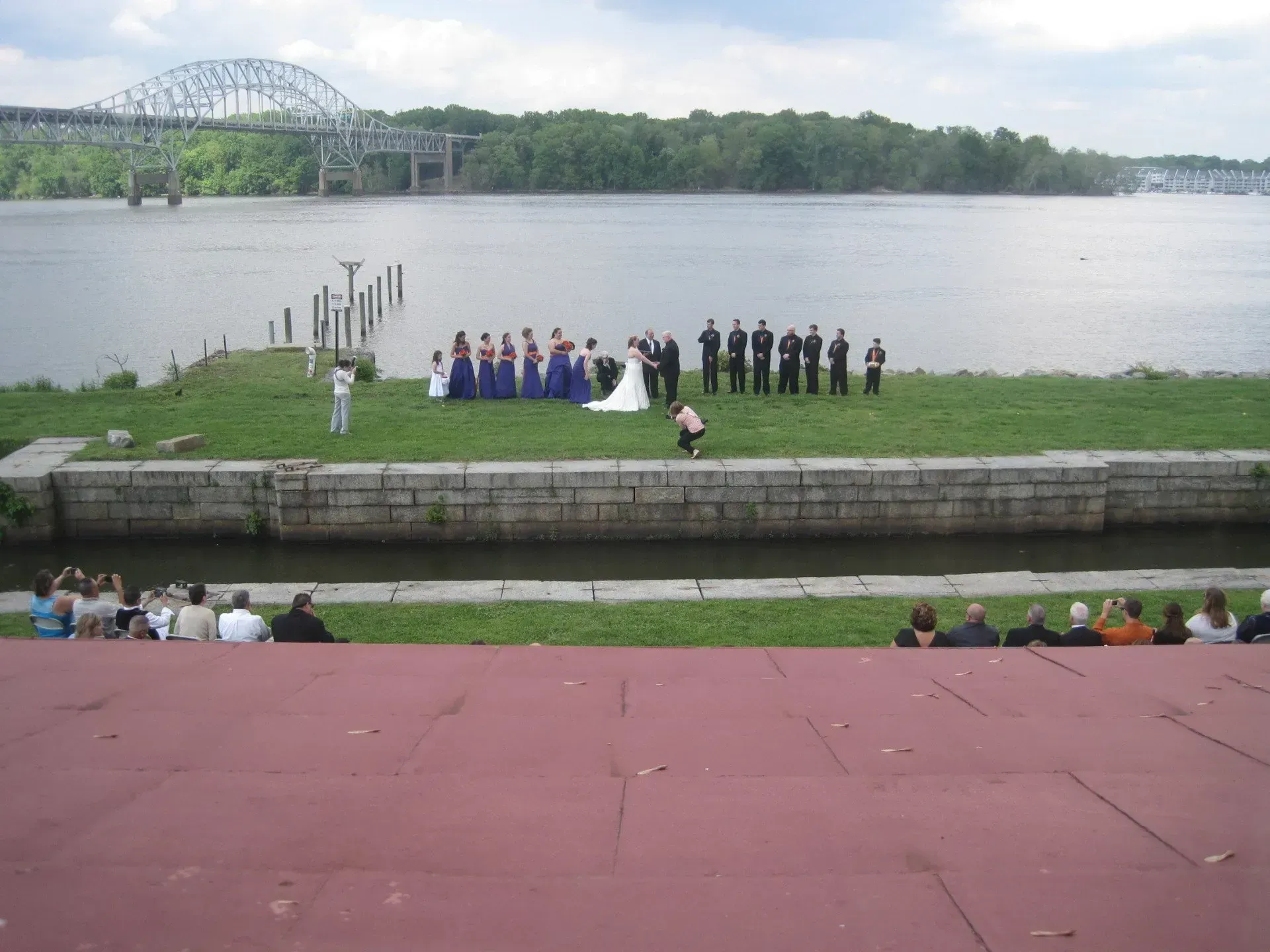 Wedding ceremony on grassy area by river with bridge in the background. Guests seated in tiered seating.