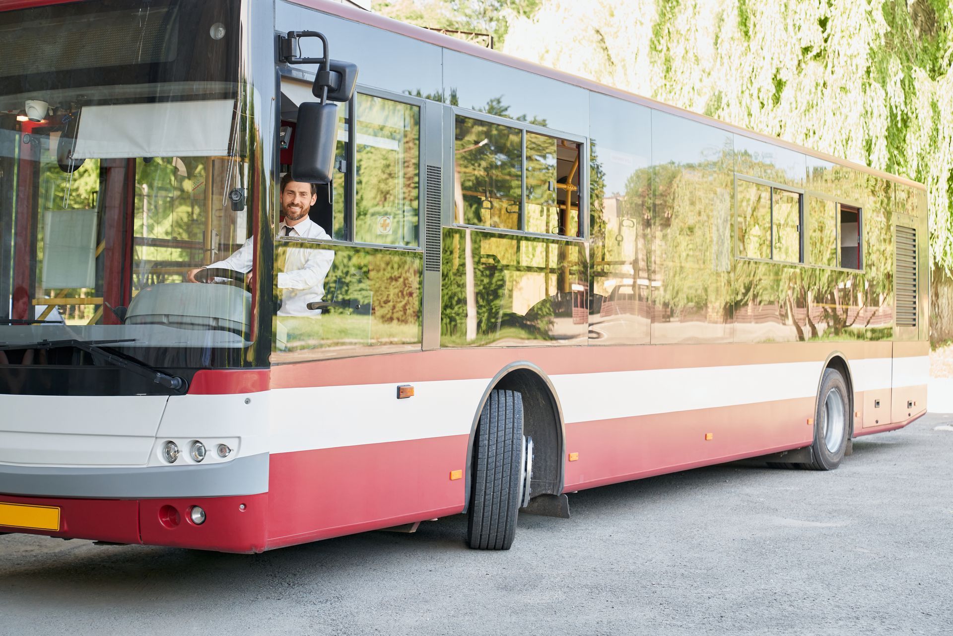 Bus driver smiling in a red and white bus. The bus is parked outdoors on a gray surface.