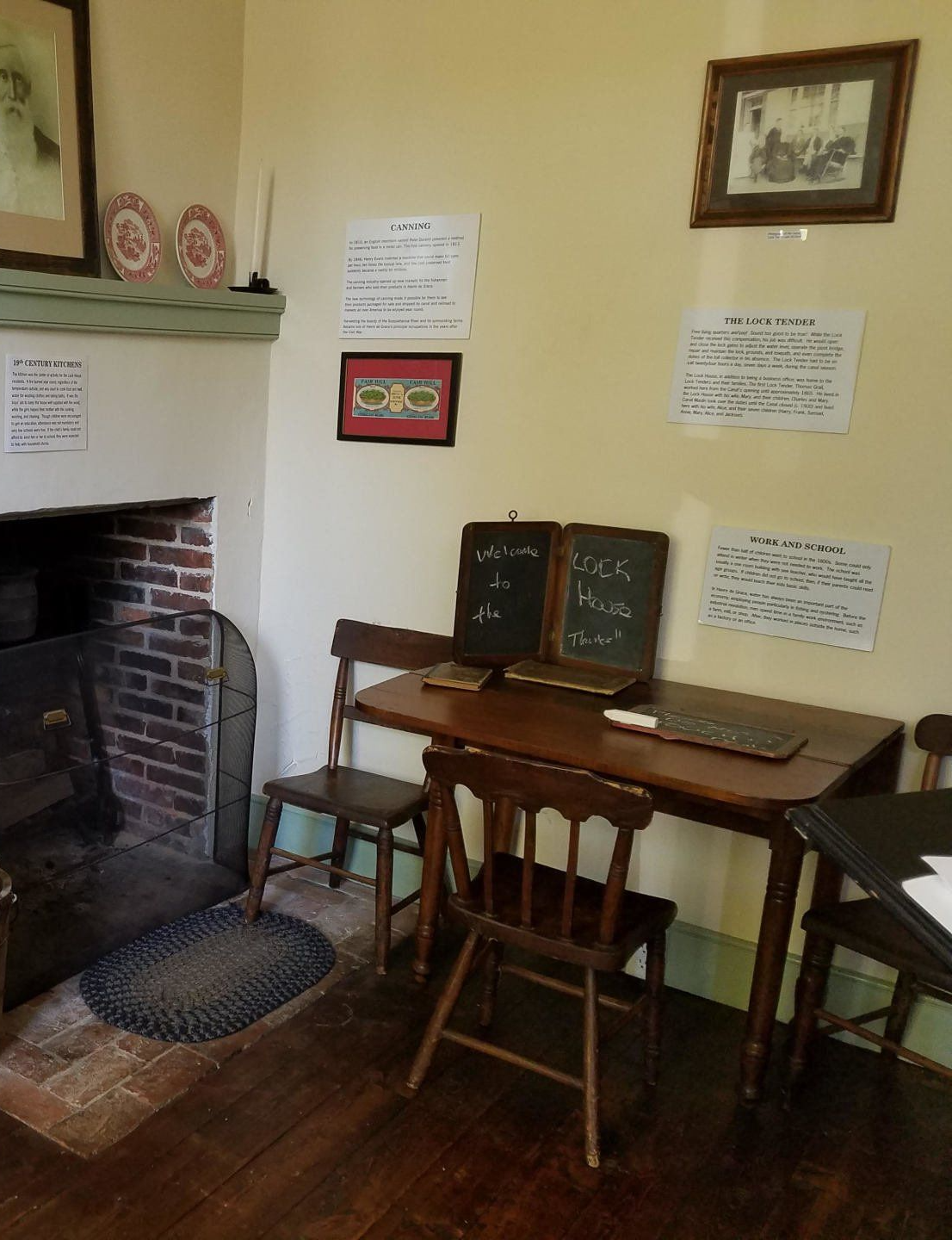 Interior of an old schoolhouse. Desk with slates, chairs, fireplace, and signs on the wall.