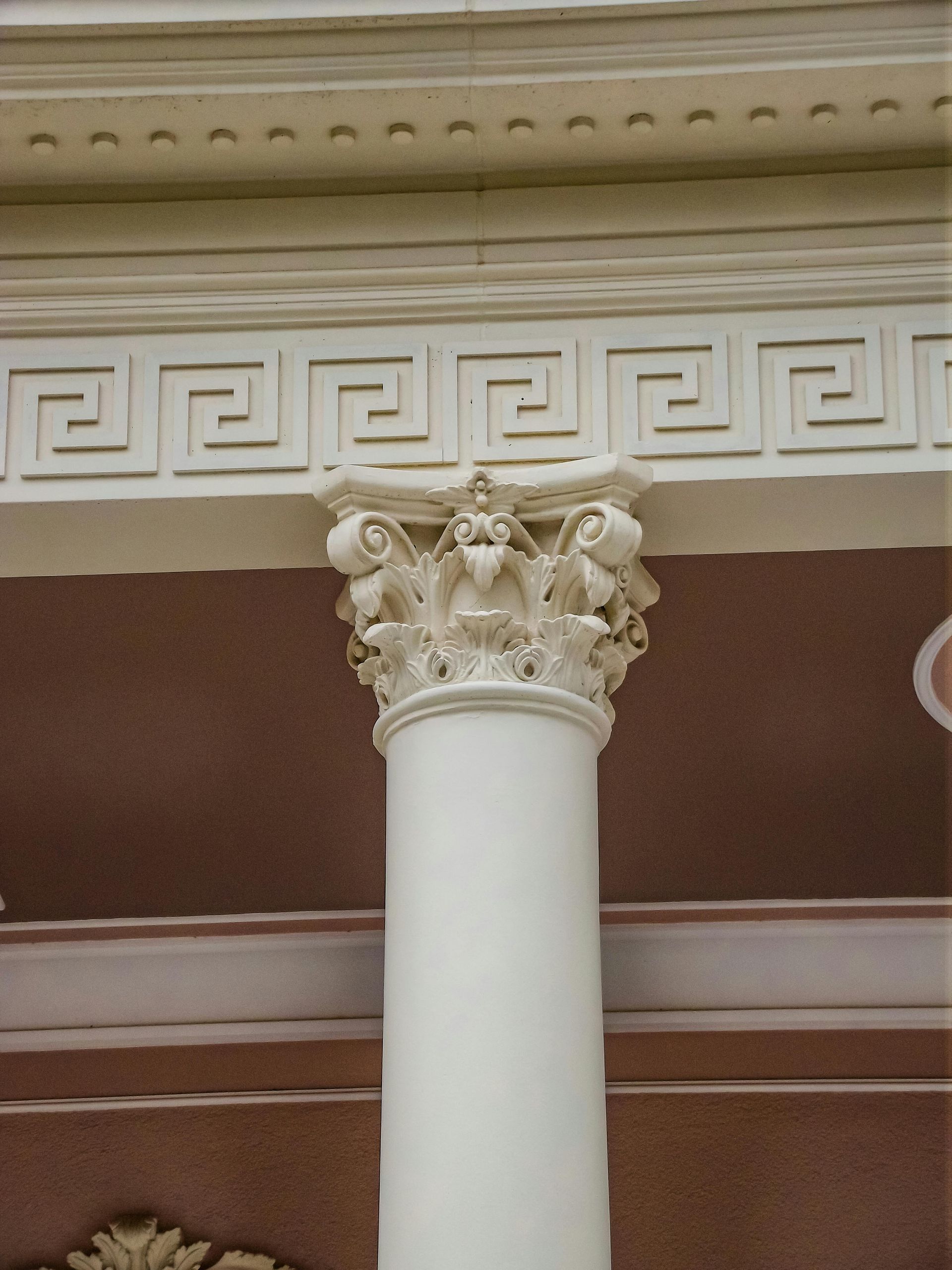 White Corinthian column topped with ornate carvings, supporting a decorative frieze with a Greek key pattern, against a pink wall.