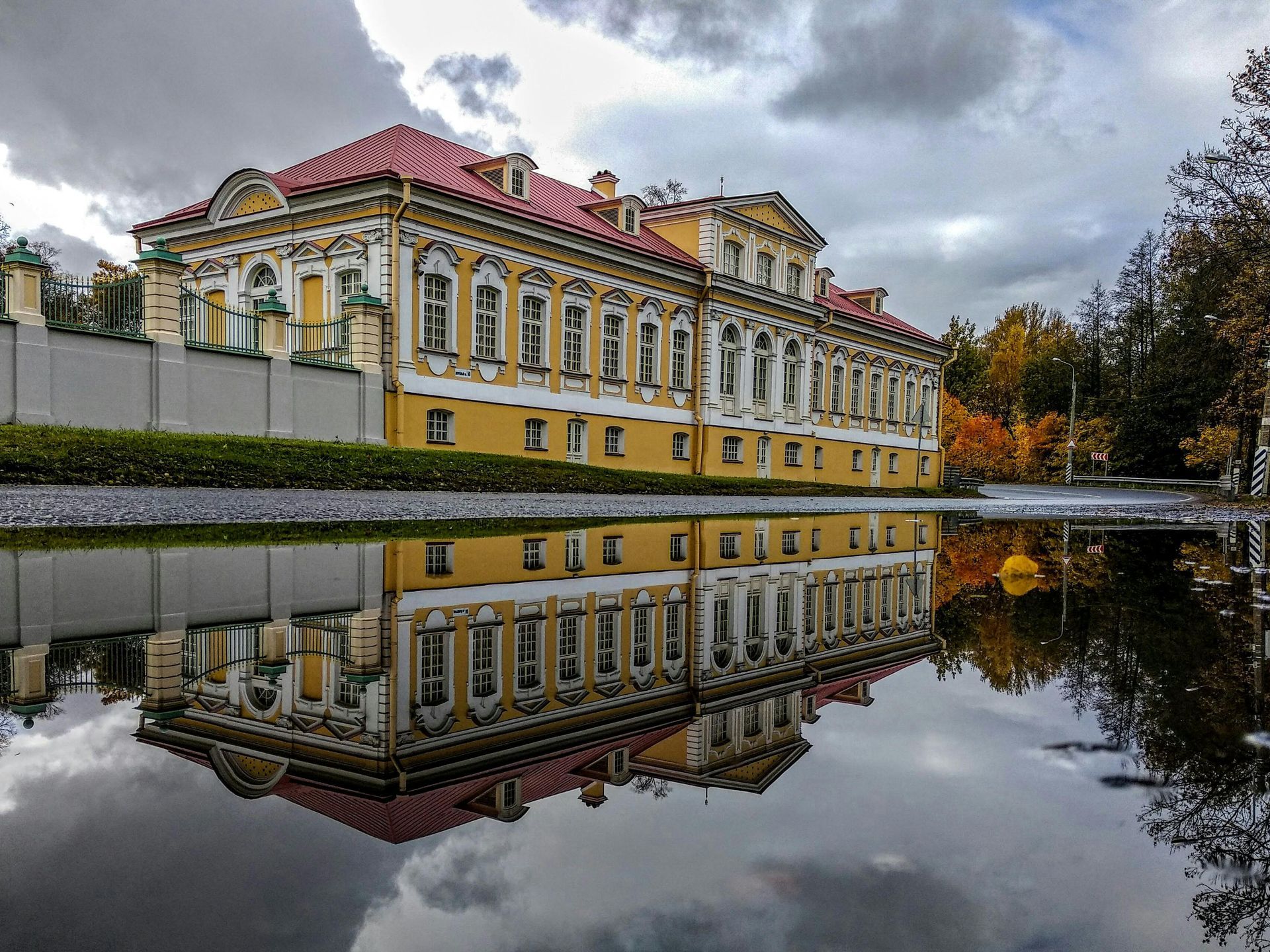 Yellow palace with red roof reflected in a puddle under a cloudy sky.