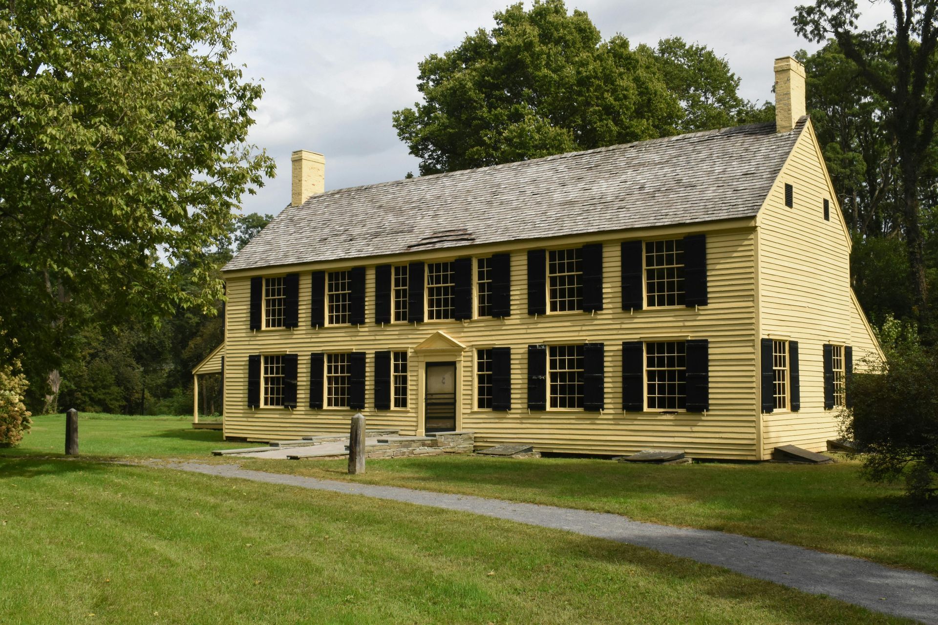 Yellow two-story house with black shutters on a grassy lawn. A paved path leads to the front door.