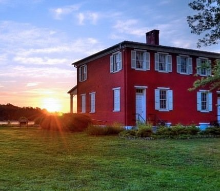 Red two-story house with white shutters, set against a sunset with a grassy lawn.