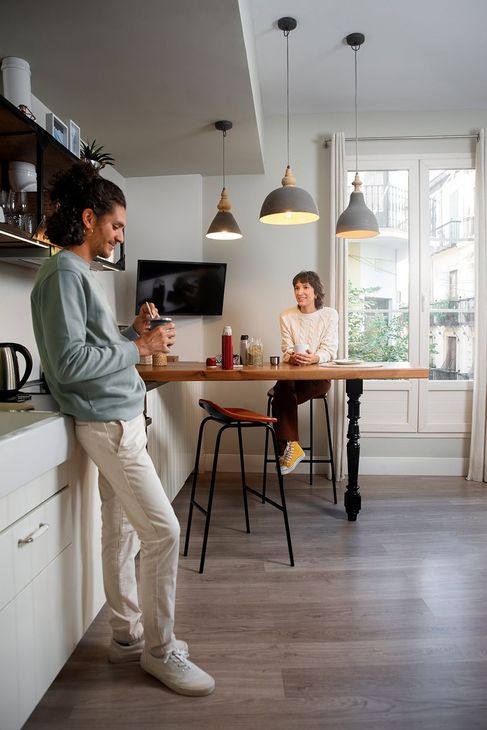 A man is leaning against a counter in a kitchen while a woman sits at a table.