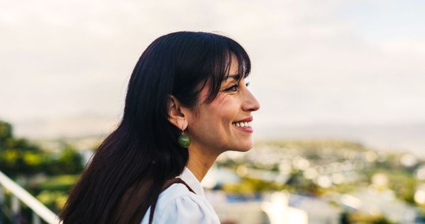 Woman with dark hair smiles, looking to the side with a city landscape backdrop.
