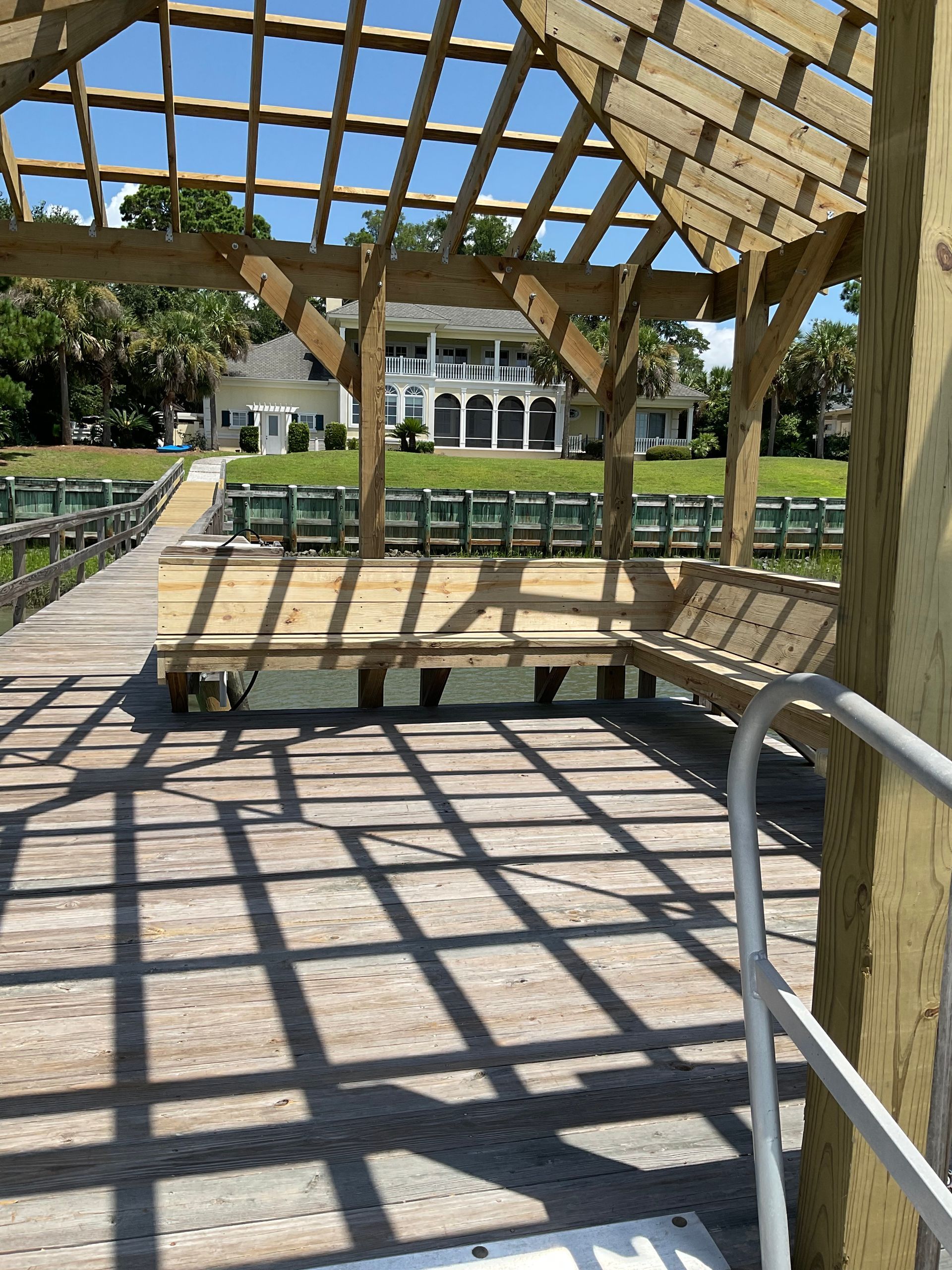A wooden pergola over a dock with a house in the background.