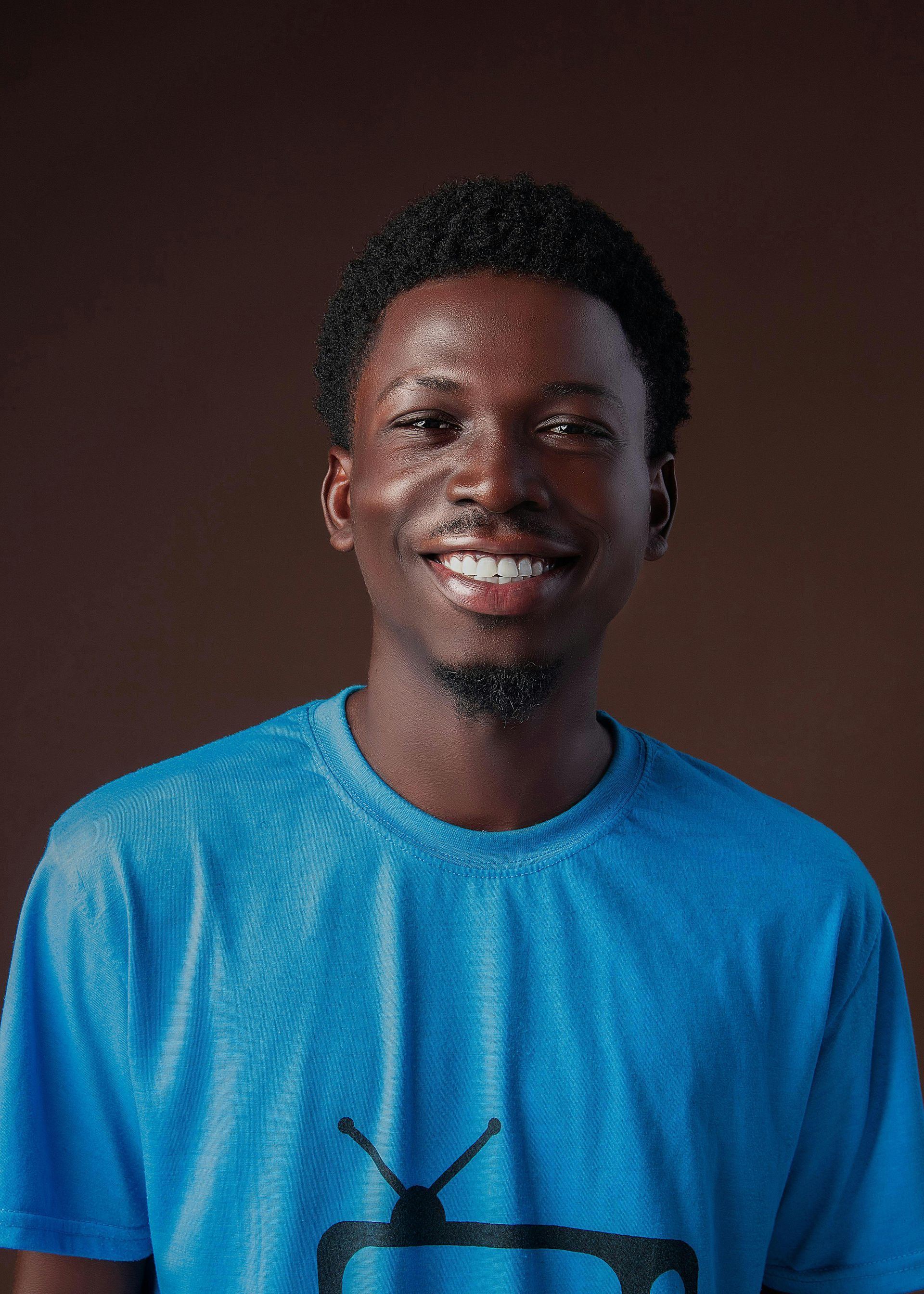 Man smiling, wearing a blue t-shirt with a TV logo, standing in front of a brown background.