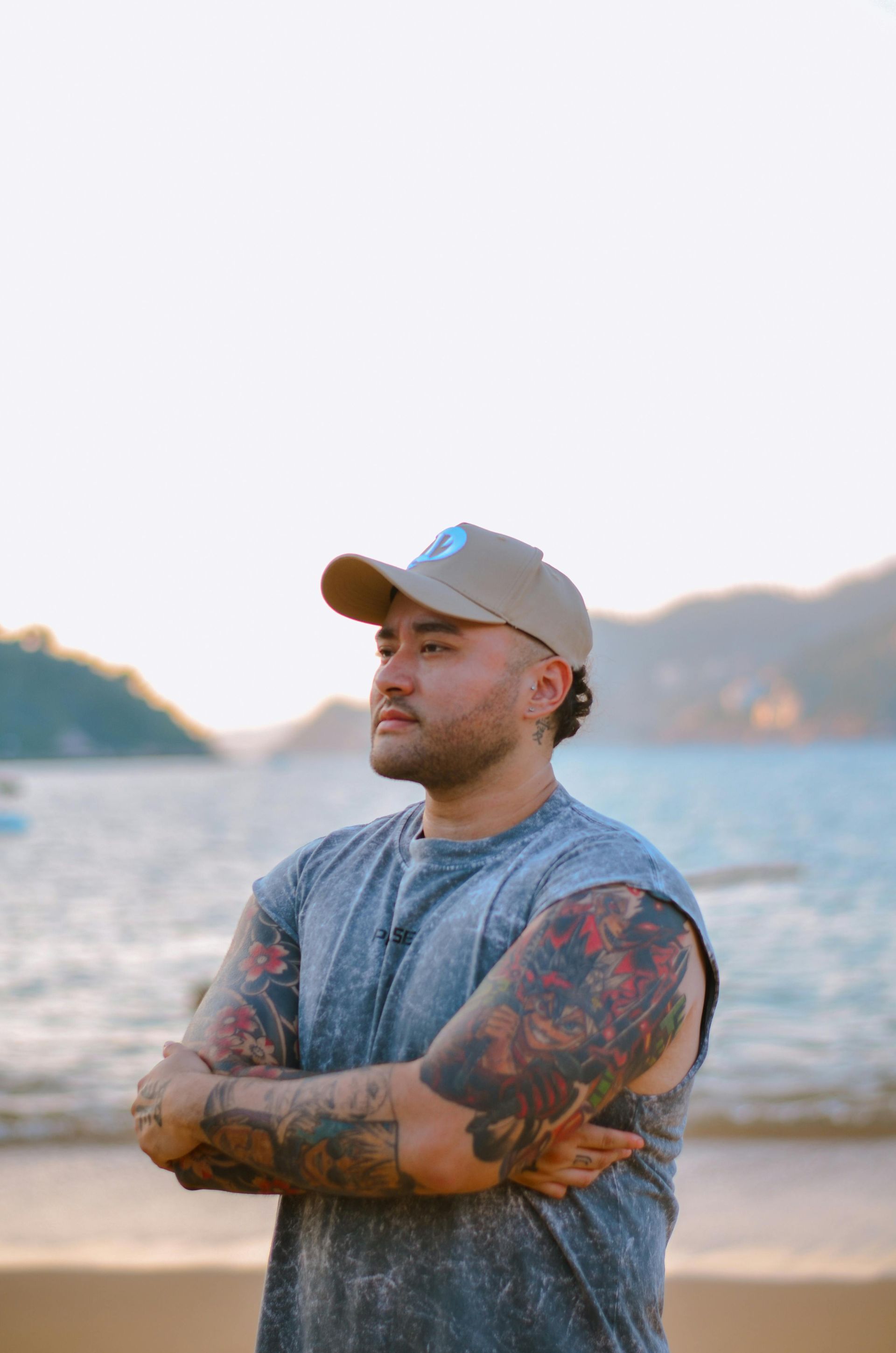 Man with tattoos and a cap, arms crossed, looking toward the ocean at sunset.