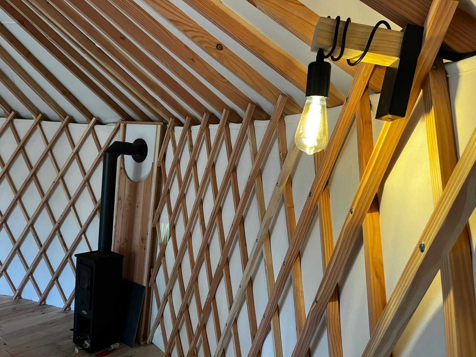Intérieur d'une yourte avec poutres en bois, un poêle noir et un luminaire suspendu.