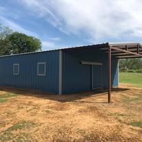 A blue metal building with a covered area underneath it in a field.