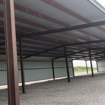A large metal building with a roof and wooden beams is sitting on top of a gravel lot.