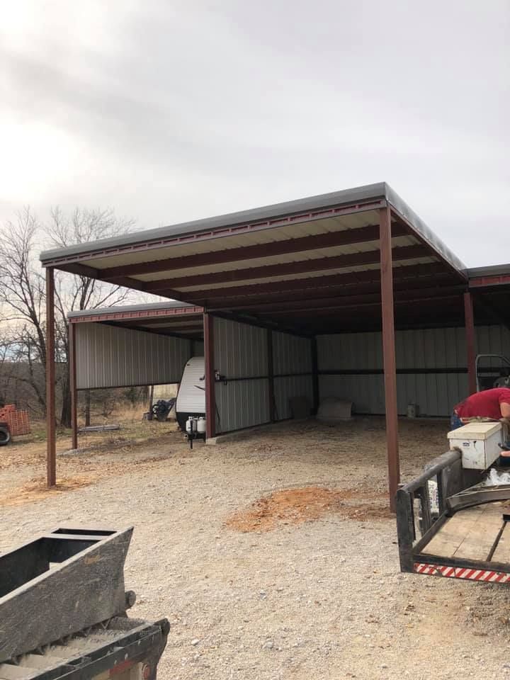 A man is standing under a covered shed in a gravel lot.