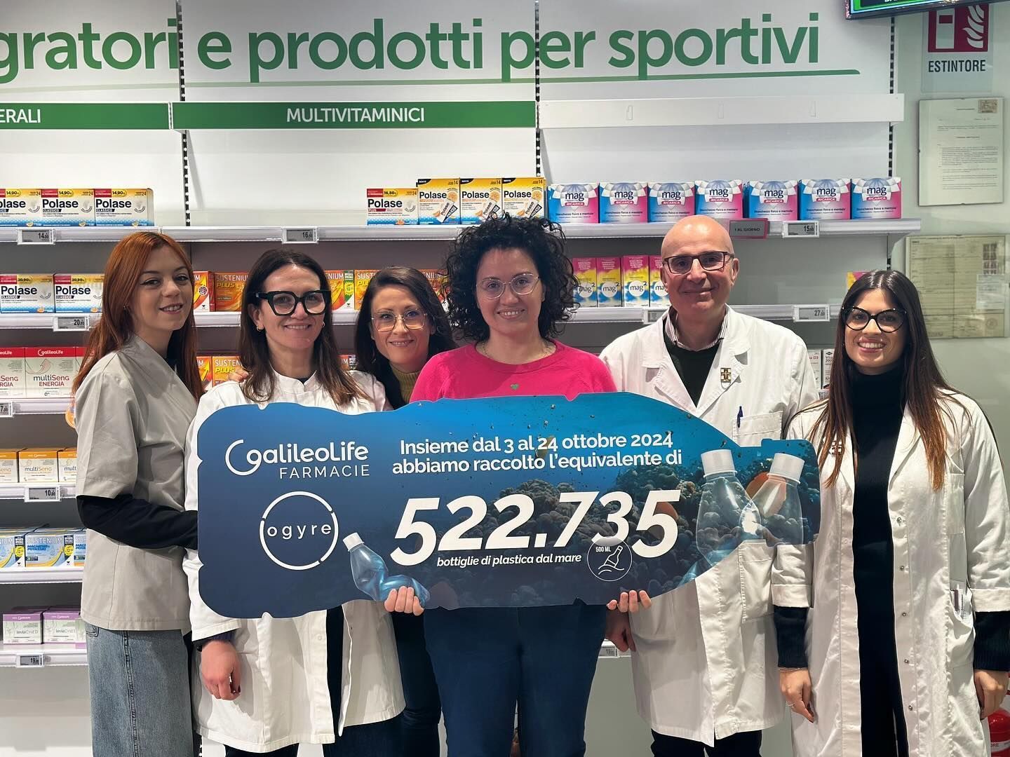 Un gruppo di persone è in piedi uno accanto all'altro in una farmacia e regge un cartello con la scritta 522 735.
