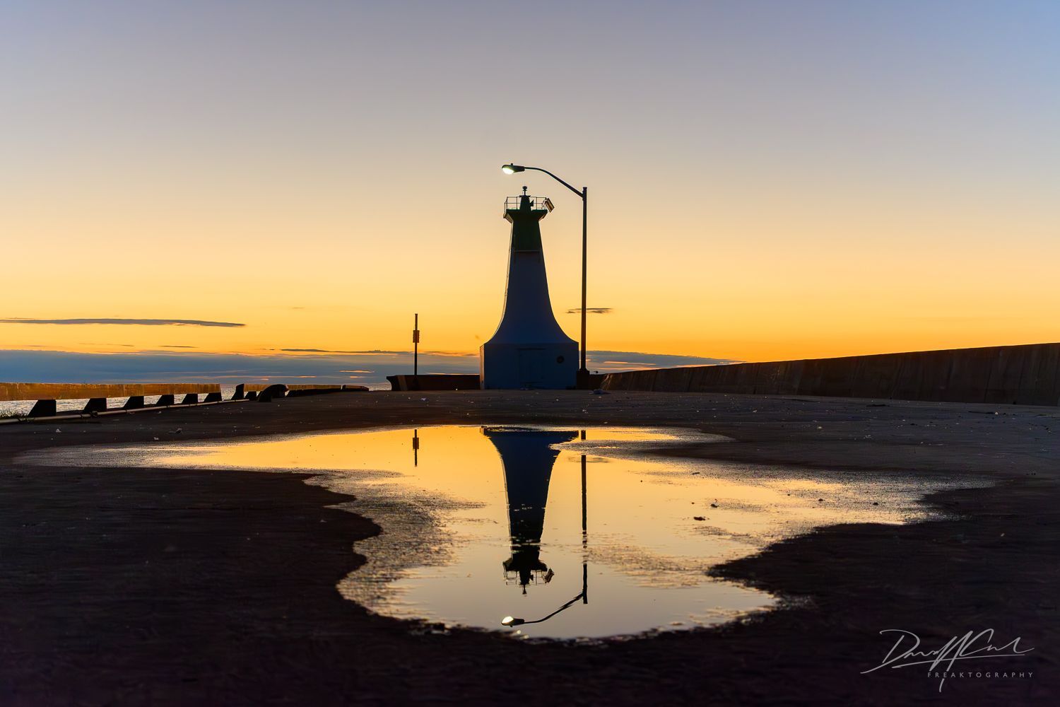 A lighthouse is reflected in a puddle of water at sunset