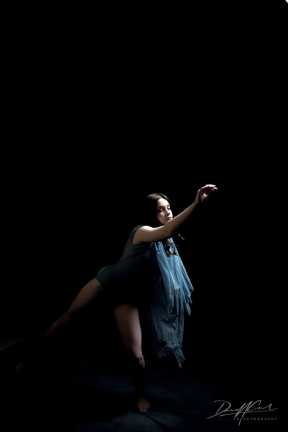 Ballerina dancing in a dark studio with one leg extended and arms reaching, lit by a single directional flash