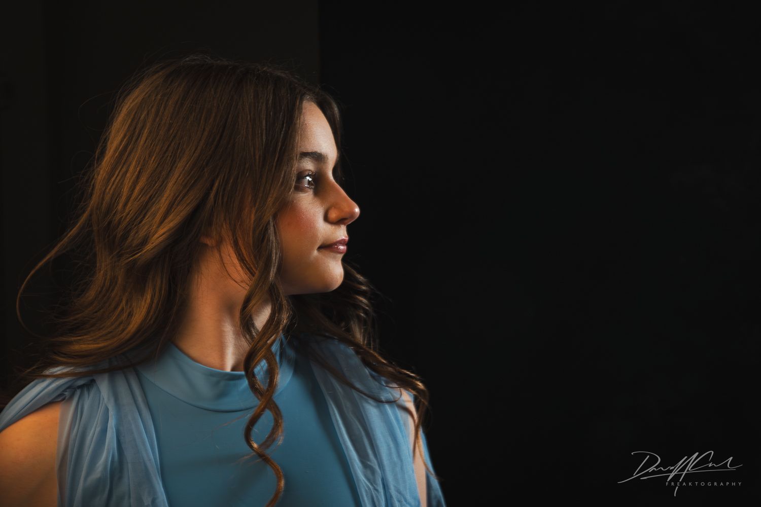 Profile portrait of a young ballerina in a soft blue costume, softly lit against a dark background