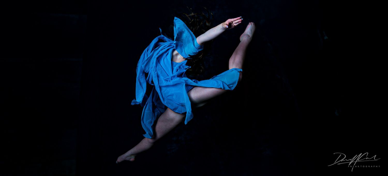 Ballerina in a blue dress frozen mid-jump in a dark studio, arms and legs extended in flight