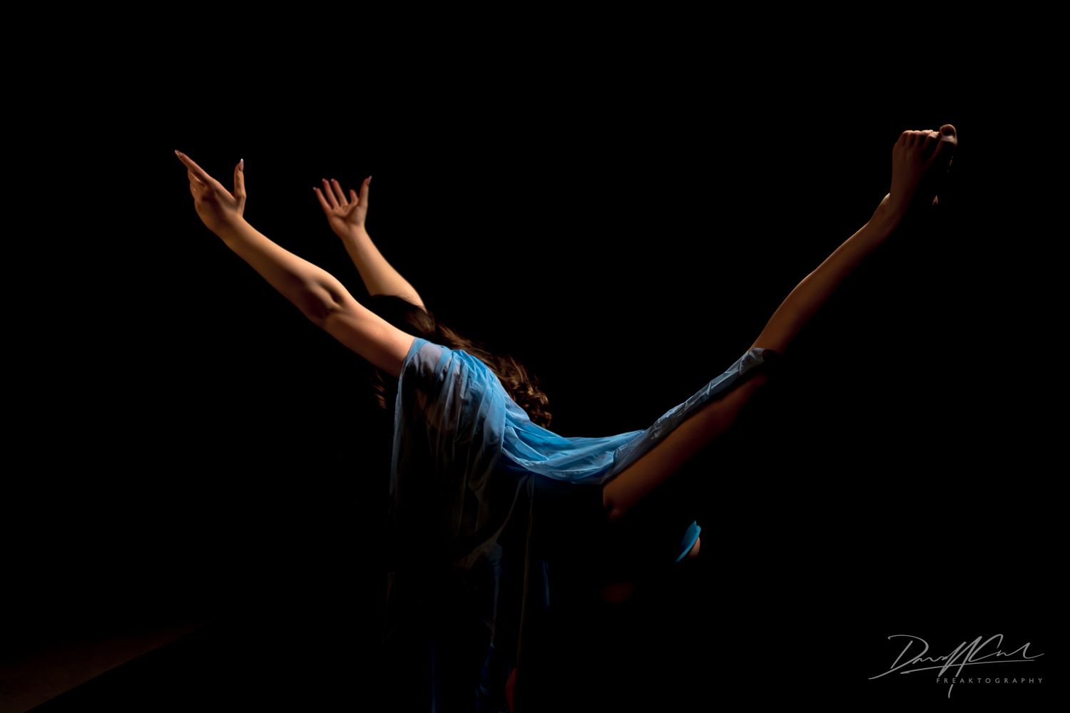 Ballerina in blue captured from behind with arms and leg extended in a dramatic pose against a black background