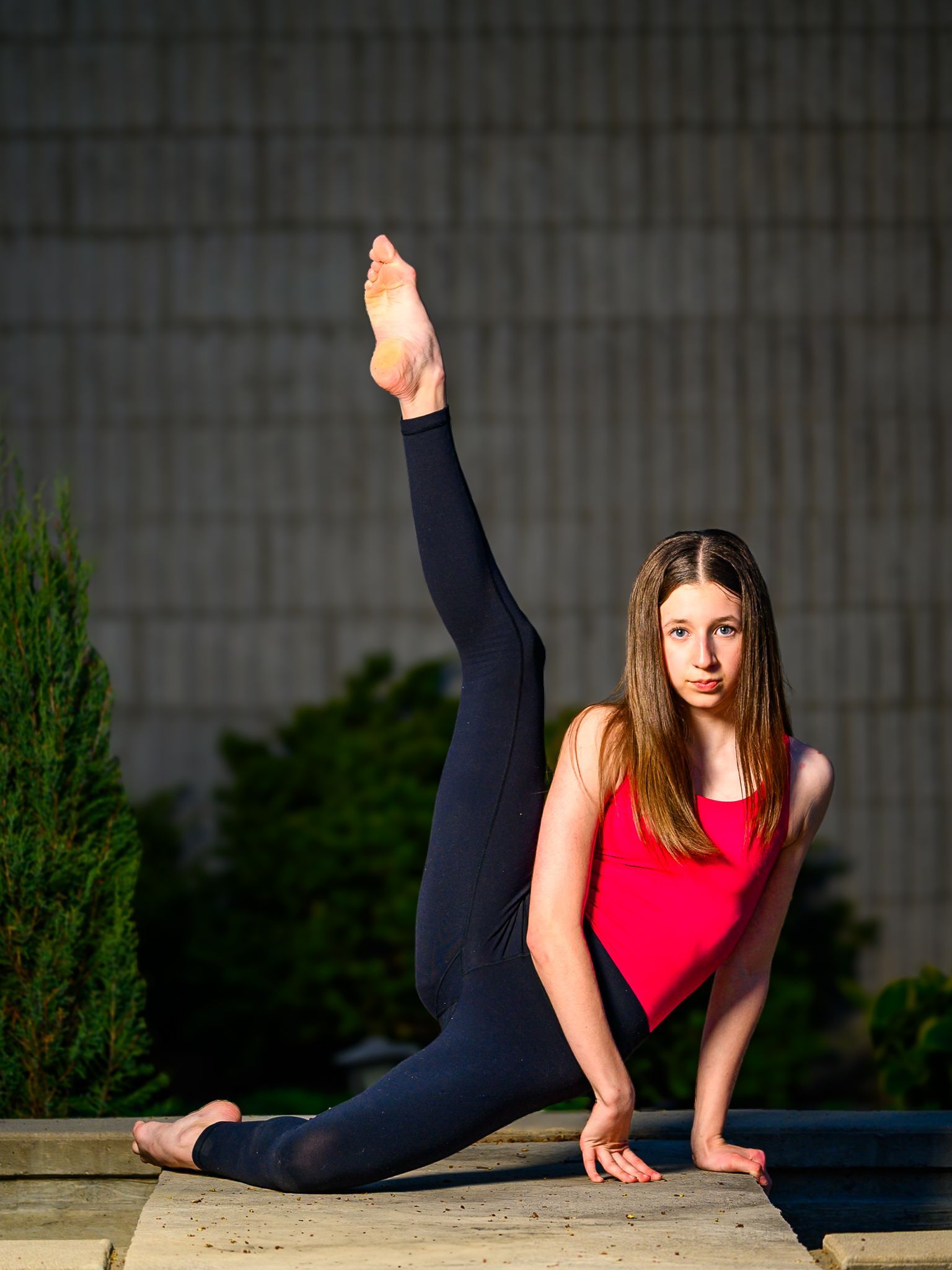 Ballerina Peyton in a red top and black leggings performing a vertical leg extension while posing on concrete outdoors