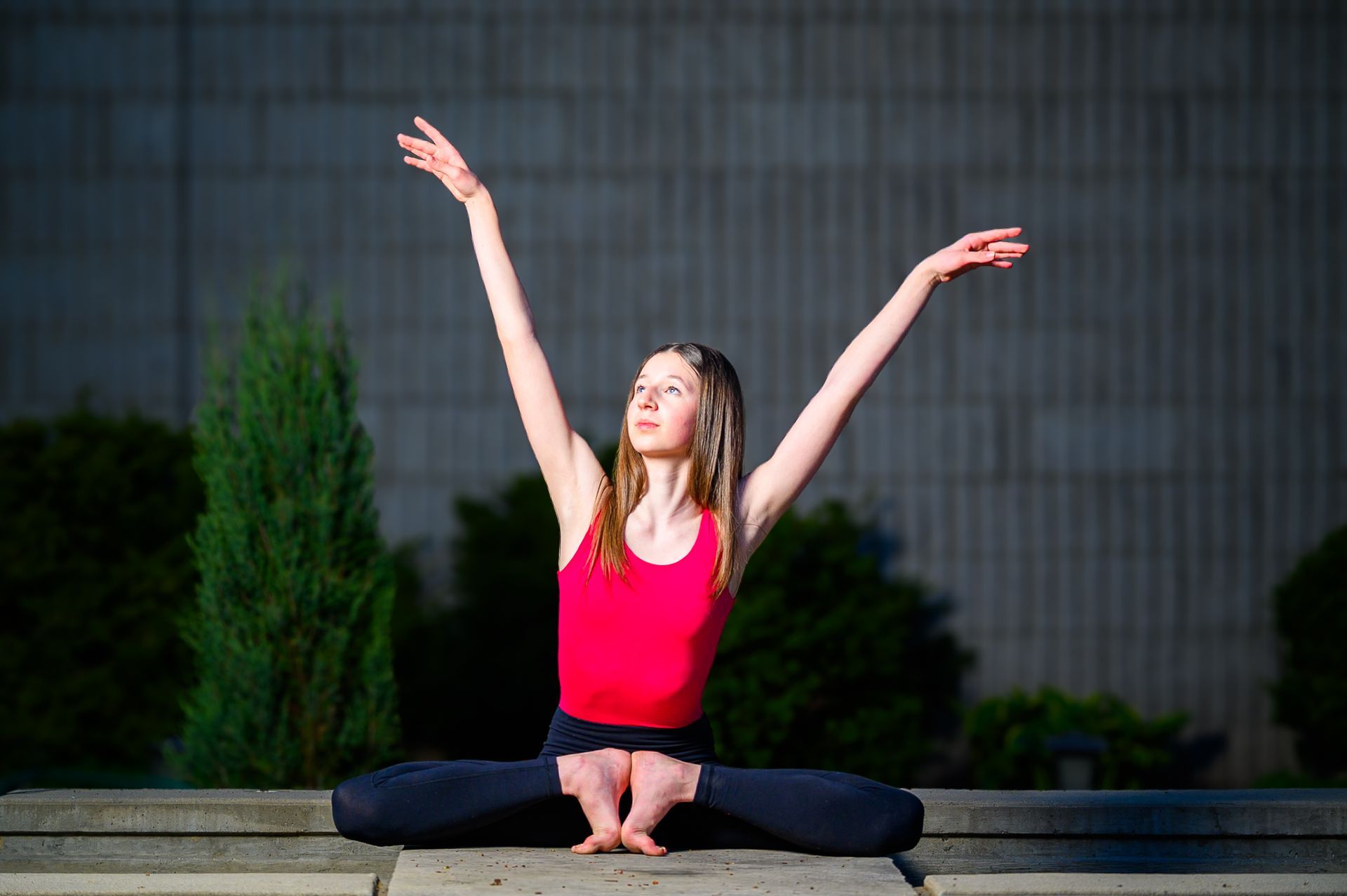 Ballerina Peyton sitting barefoot with legs crossed and arms raised upward, outdoors against a concrete wall and trees