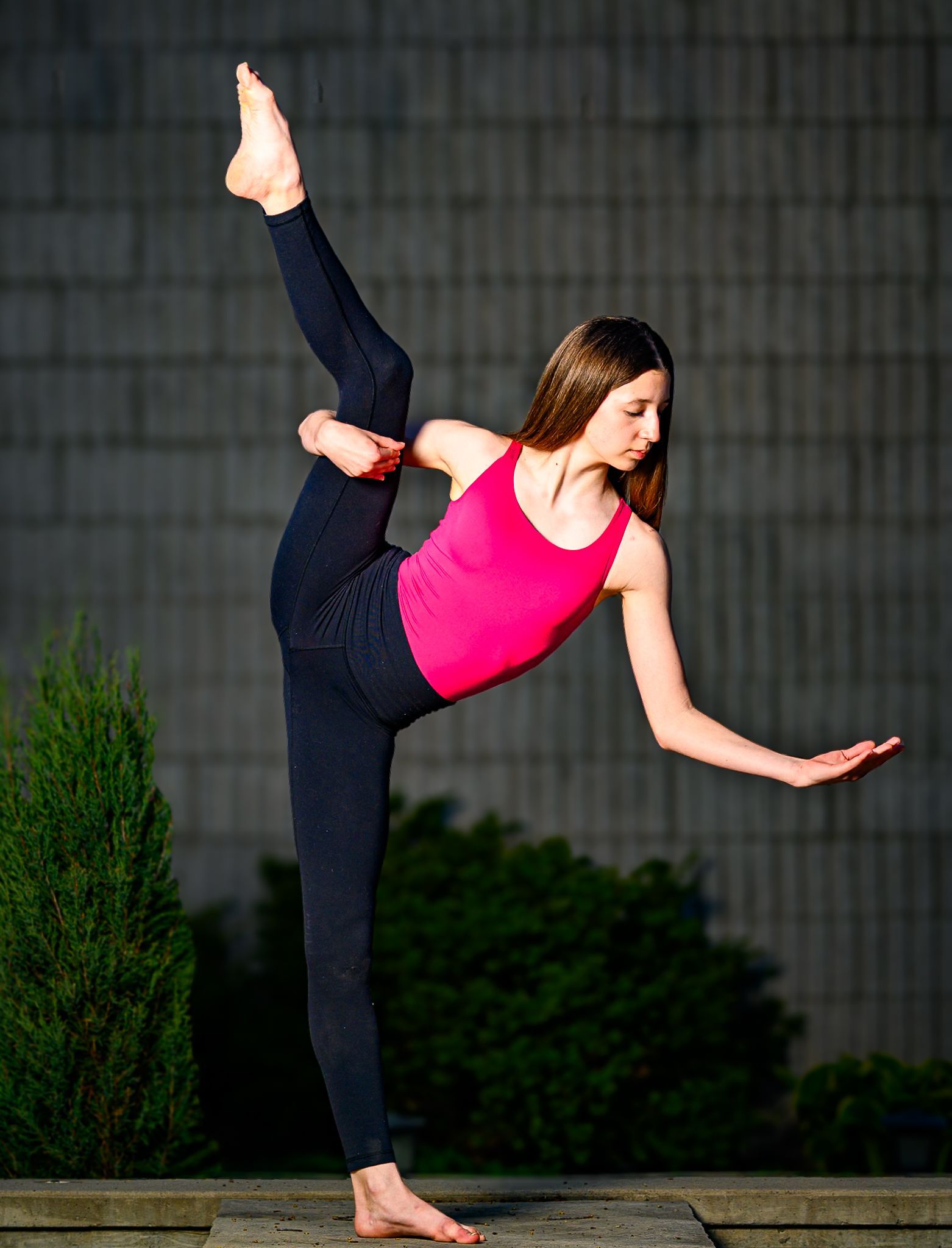 Ballerina Peyton in a red top and black leggings holding a high leg pose barefoot outdoors against a grey wall