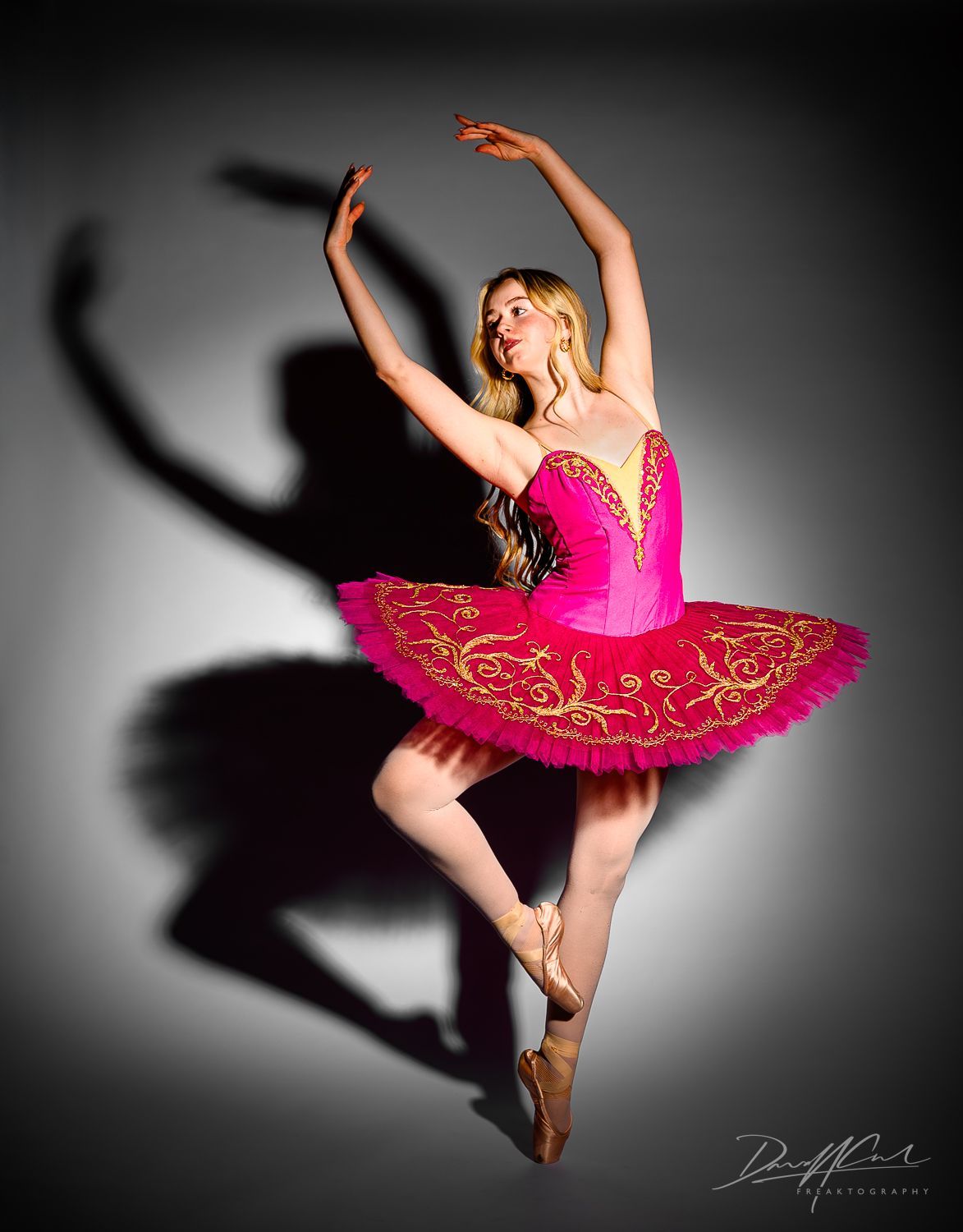 Ballet dancer en pointe in a red and gold tutu casting a dramatic shadow on a grey studio wall