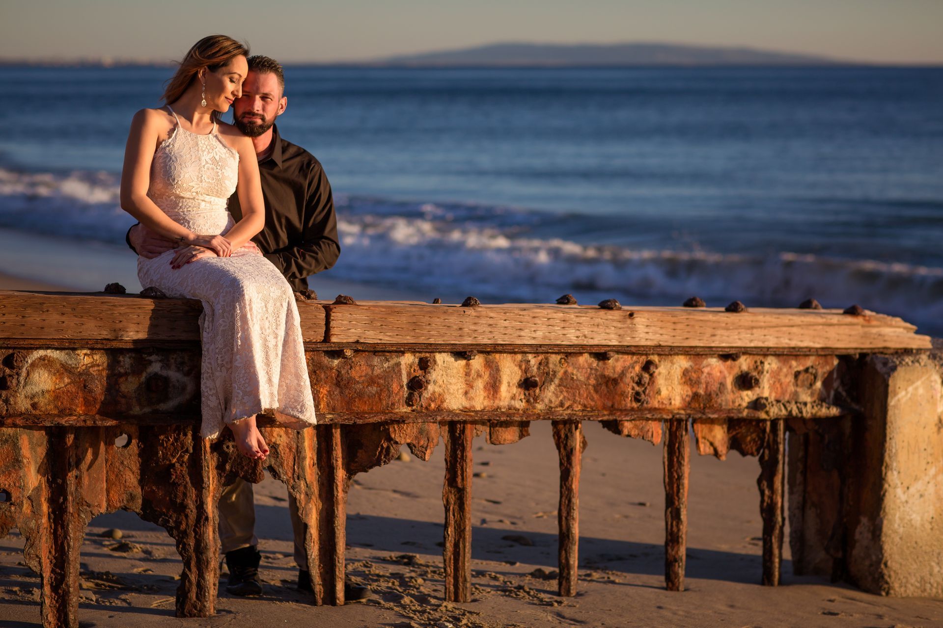 Romantic beach engagement portrait of a couple sitting on a rusted pier at sunset, with soft golden 