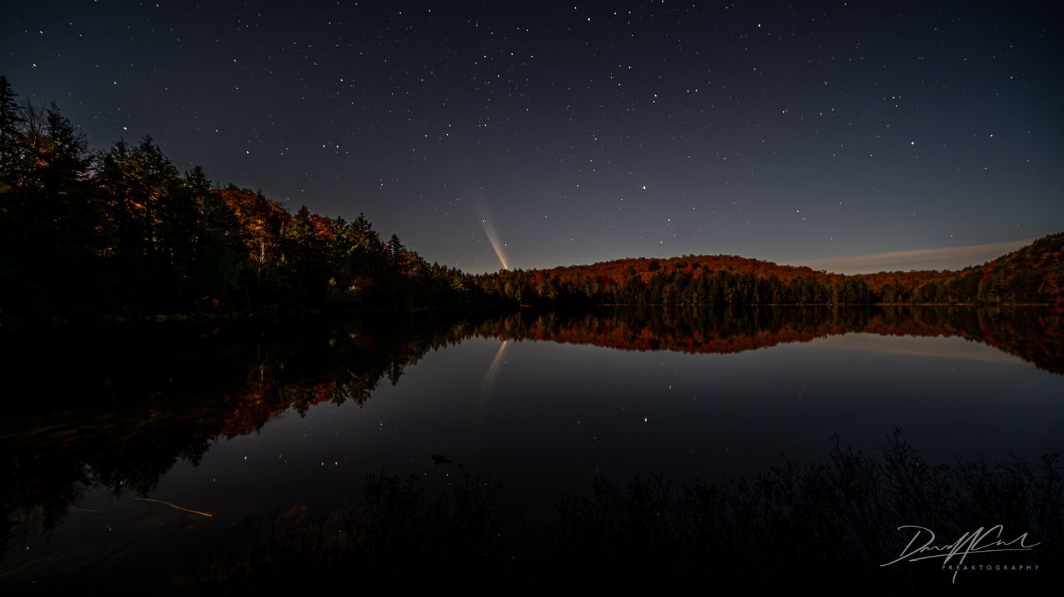 A night sky with stars over a lake with trees in the foreground.