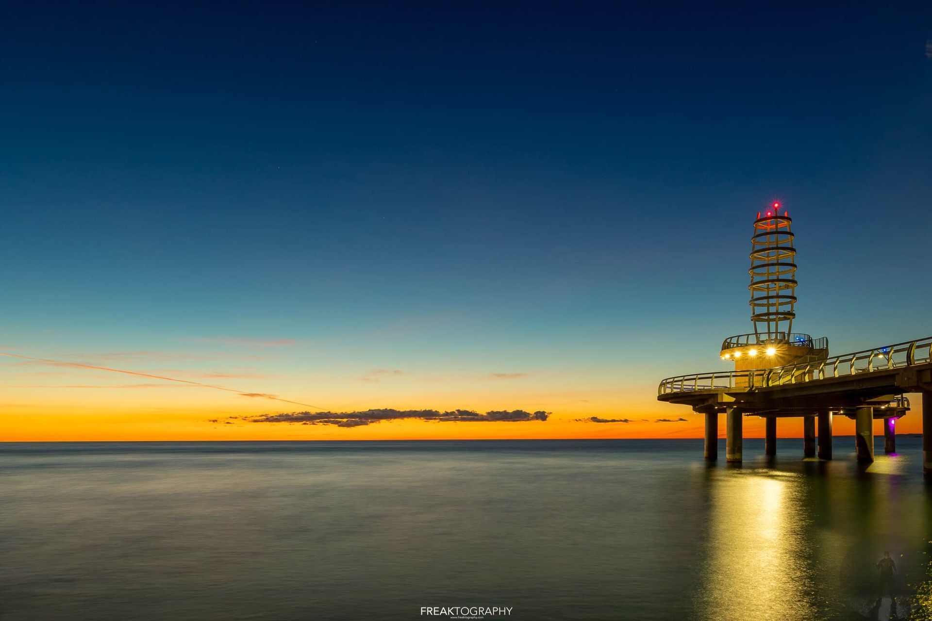 A pier overlooking the ocean at sunset with a lighthouse in the background.