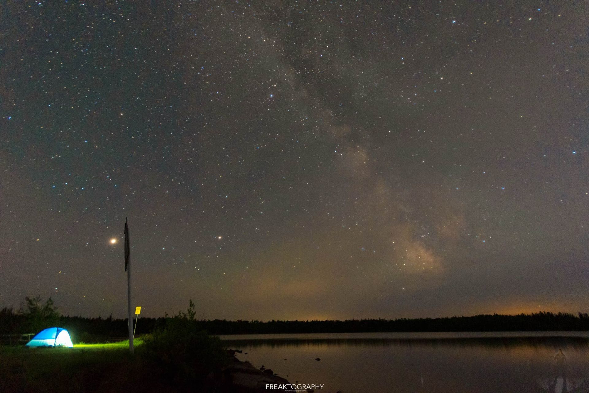 A tent is sitting on the shore of a lake under a starry night sky.