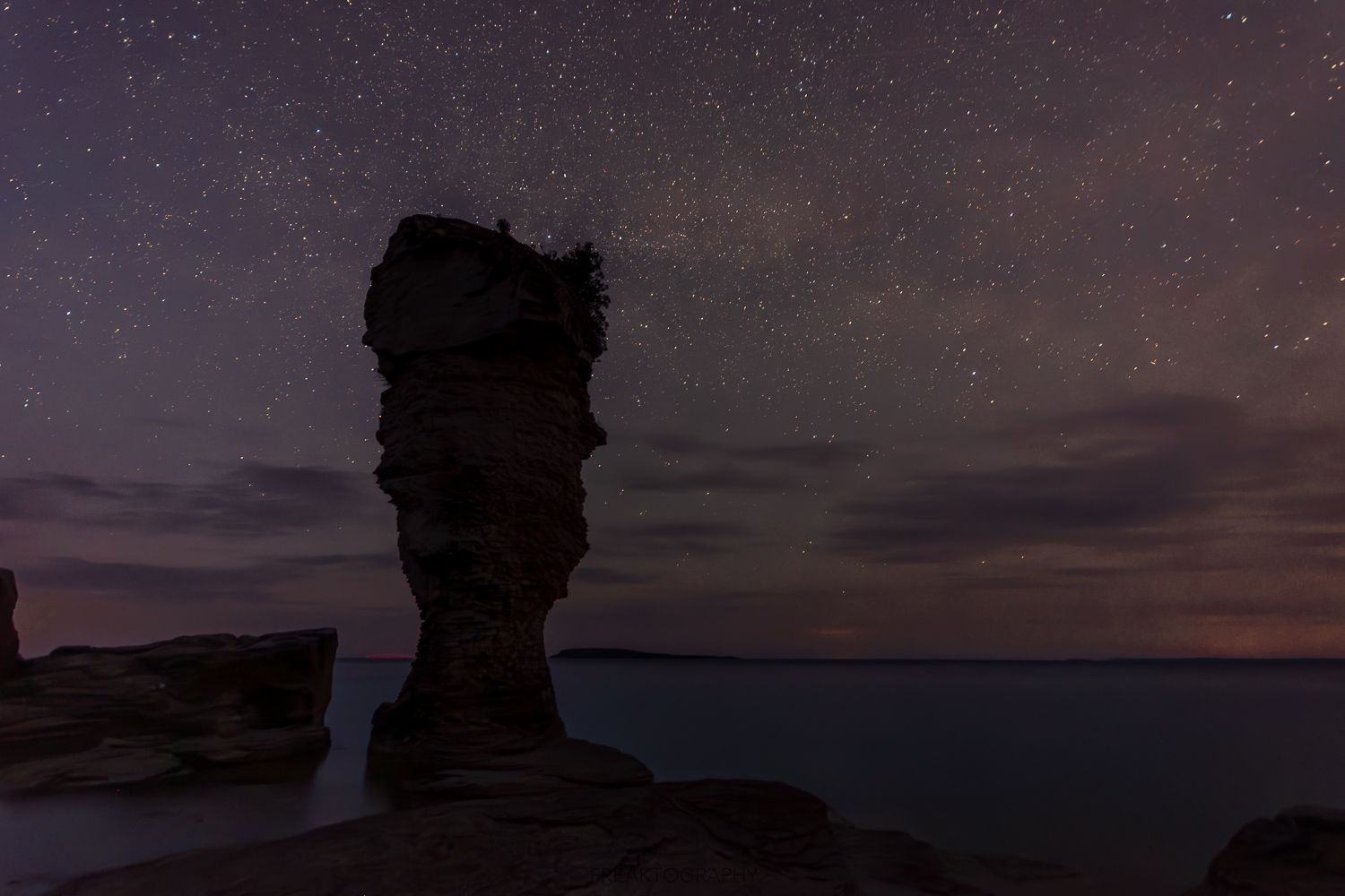 A rock formation in the middle of the ocean at night under a starry sky.