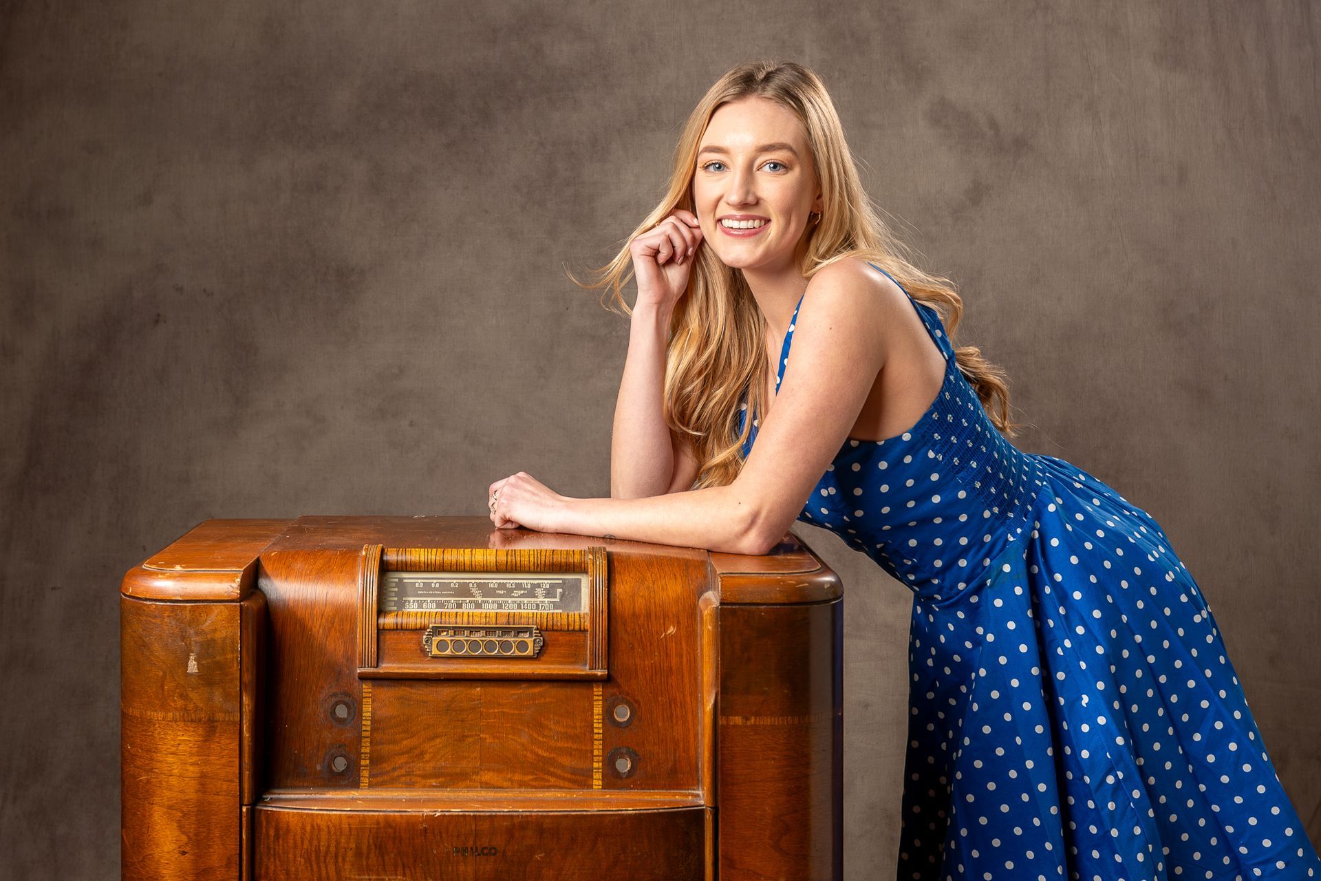 Portrait of a woman in a blue polka dot dress leaning on a vintage wooden radio against a neutral studio backdrop