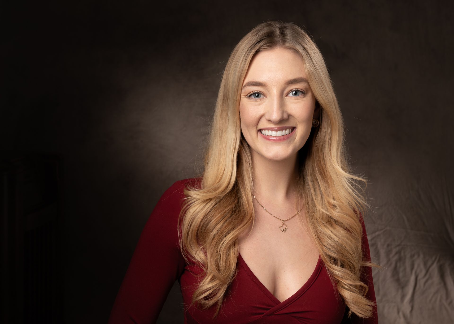 Smiling woman with long blonde hair in a red dress, posed against a brown studio backdrop