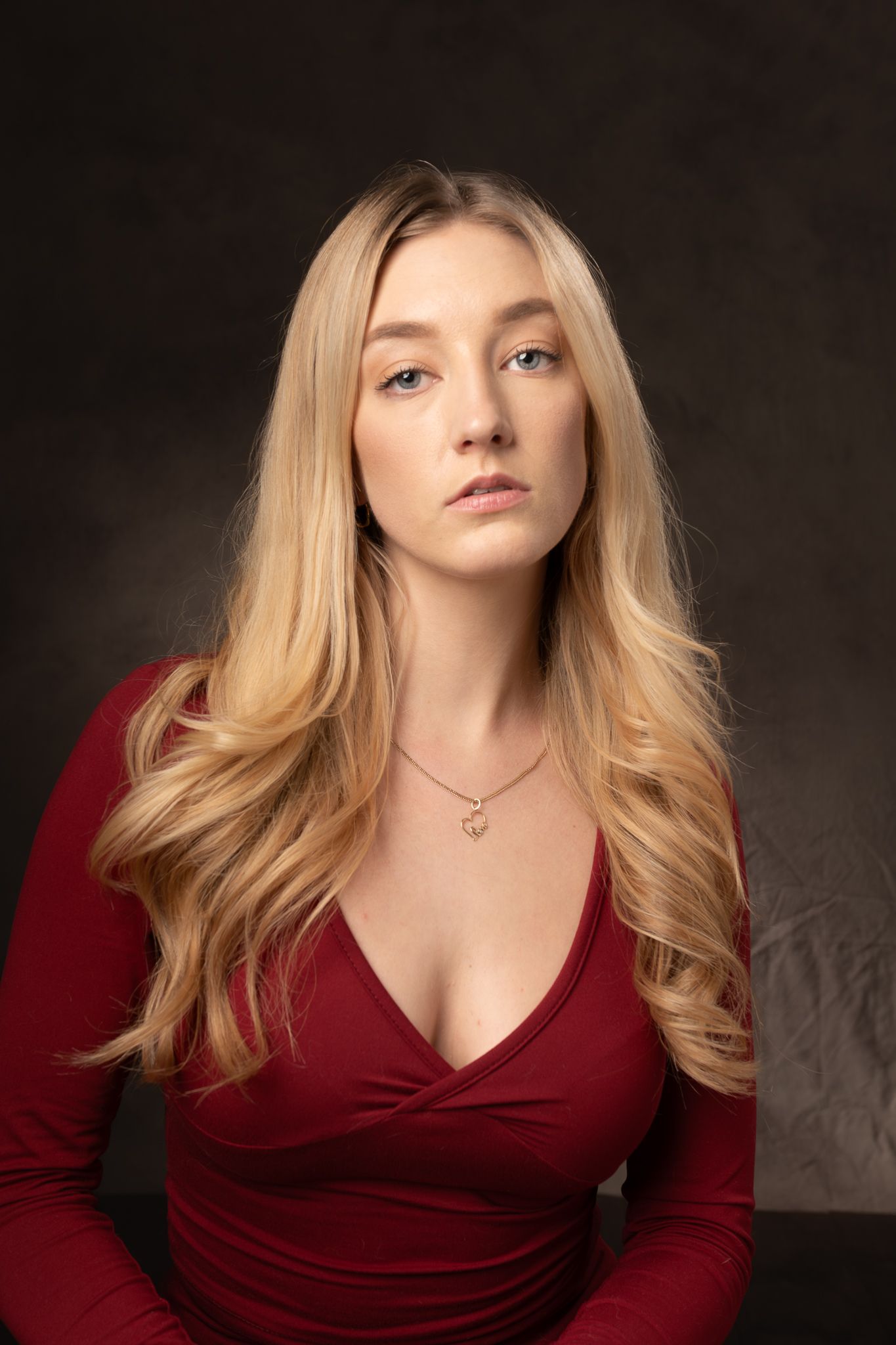 Woman with long blonde hair in a red dress sitting against a dark studio background with a neutral expression