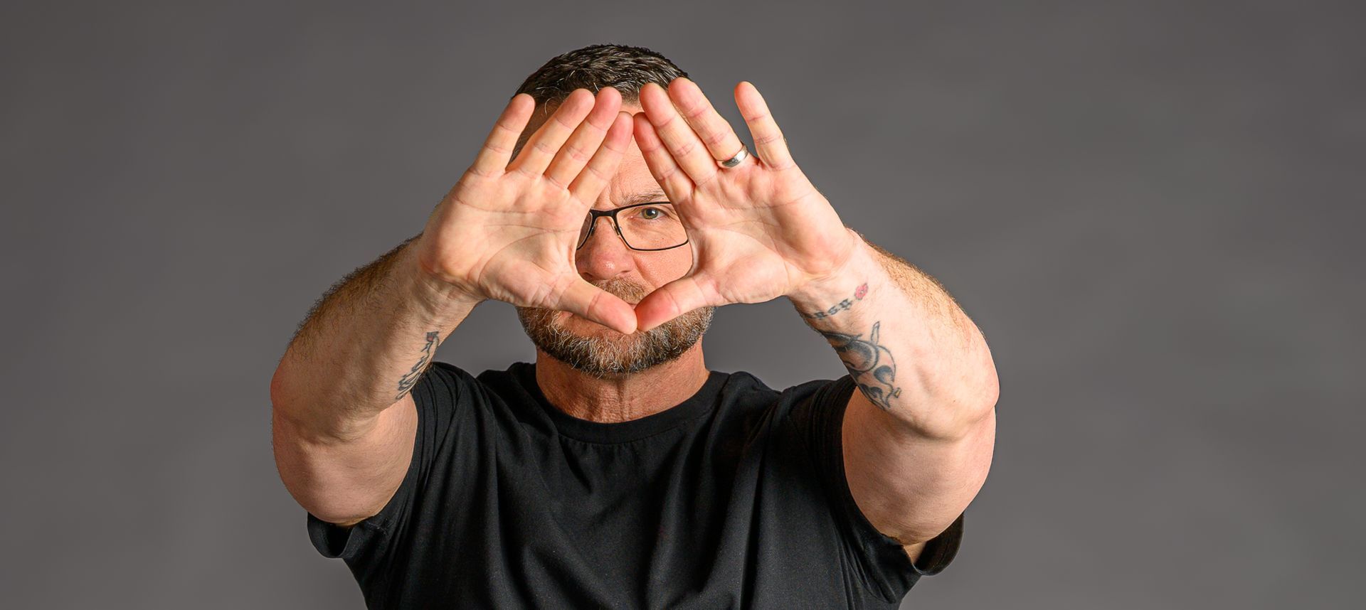 Self-portrait of Canadian photographer Dave Conlon making a triangle shape with his hands in front of his face, tattoos visible on his arms