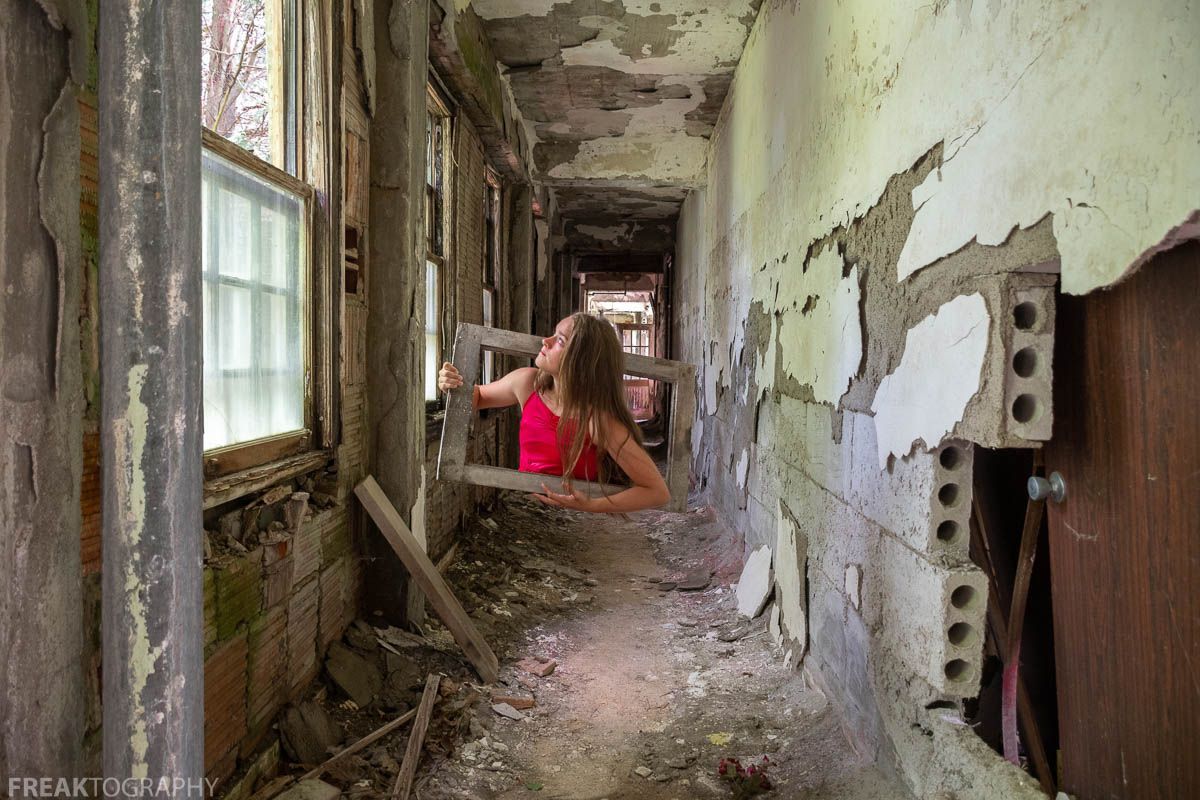 Portrait of a girl in a red top appearing to climb through a wooden frame inside an abandoned hallway with peeling paint
