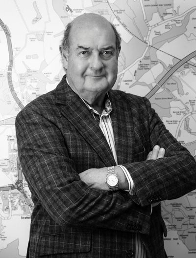 A man in a plaid suit is standing with his arms crossed in front of a map.