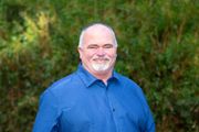 A man in a blue shirt is standing in front of a bush.