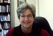 A woman wearing glasses is smiling while sitting in a chair in front of a bookshelf.