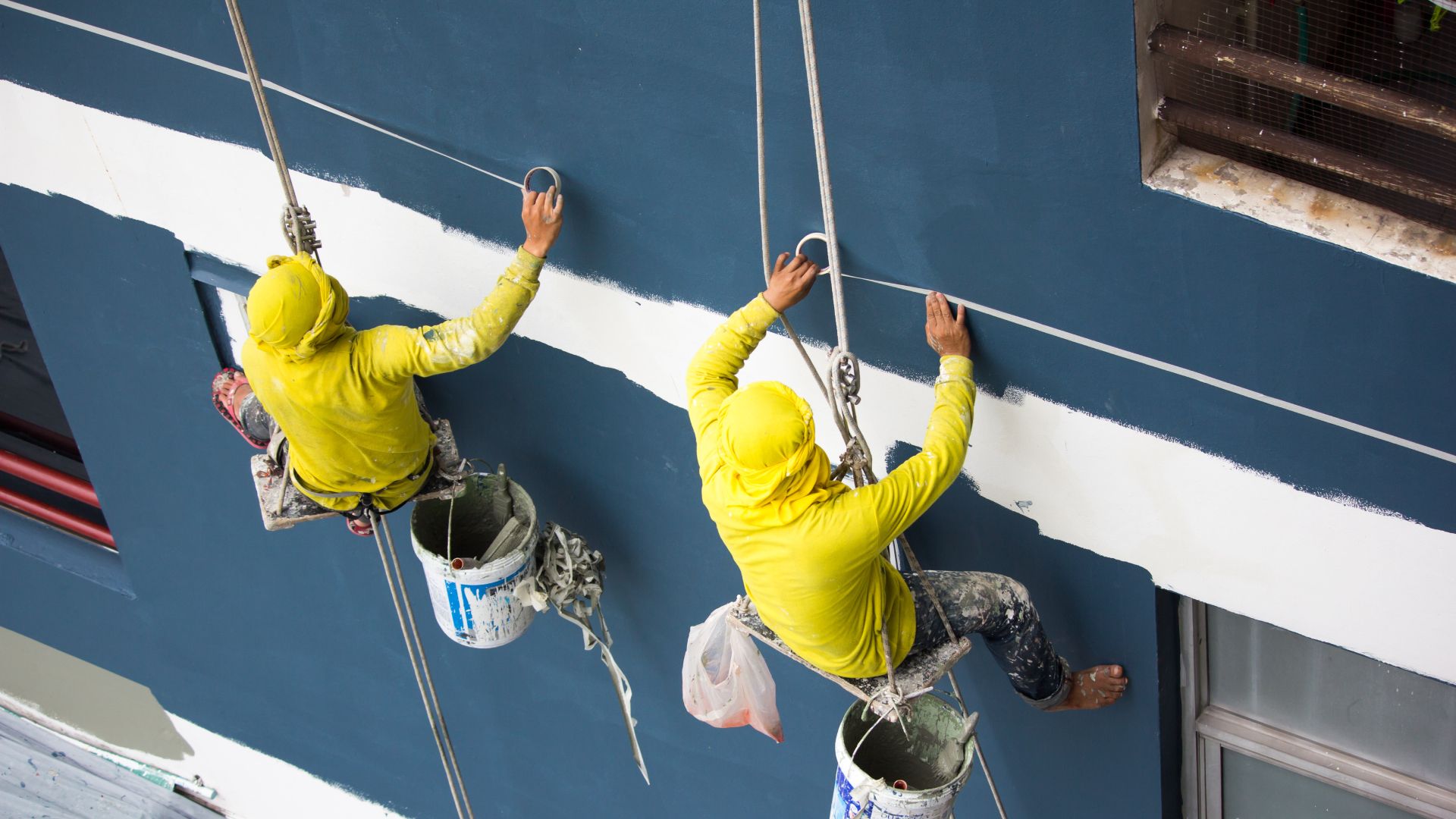 Two men are painting a building with buckets attached to ropes.