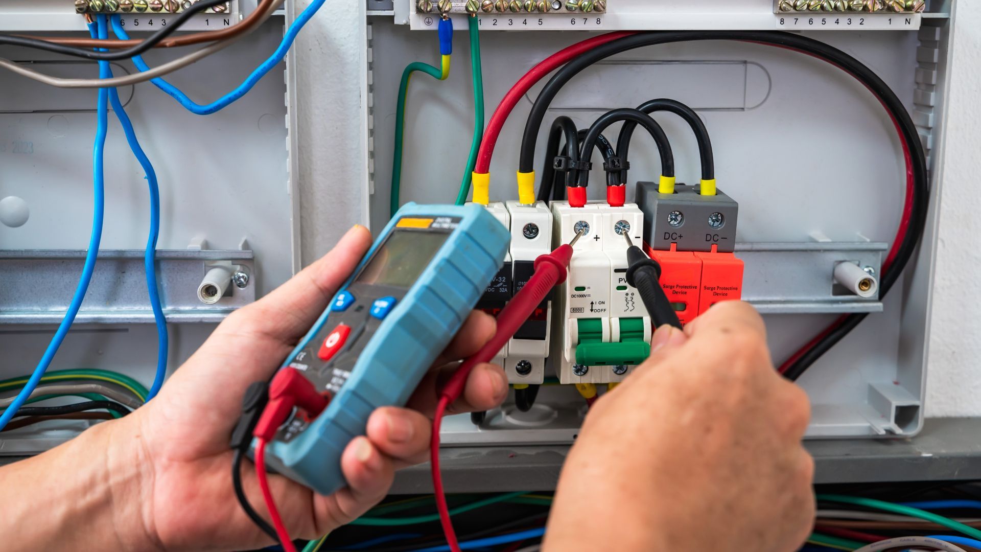 A person is holding a multimeter in their hand in front of an electrical box.