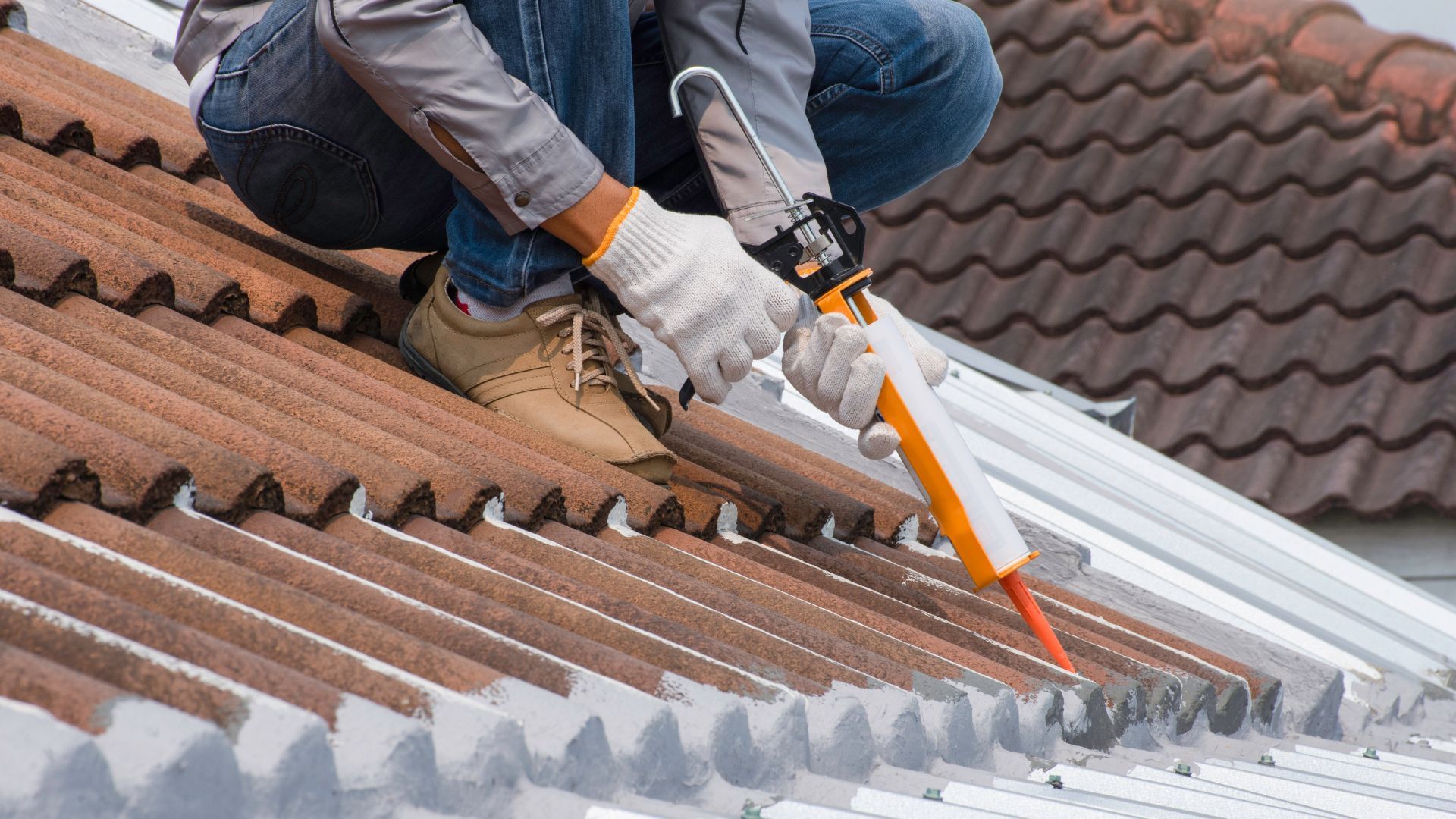 A man is kneeling on the roof of a building applying silicone sealant.