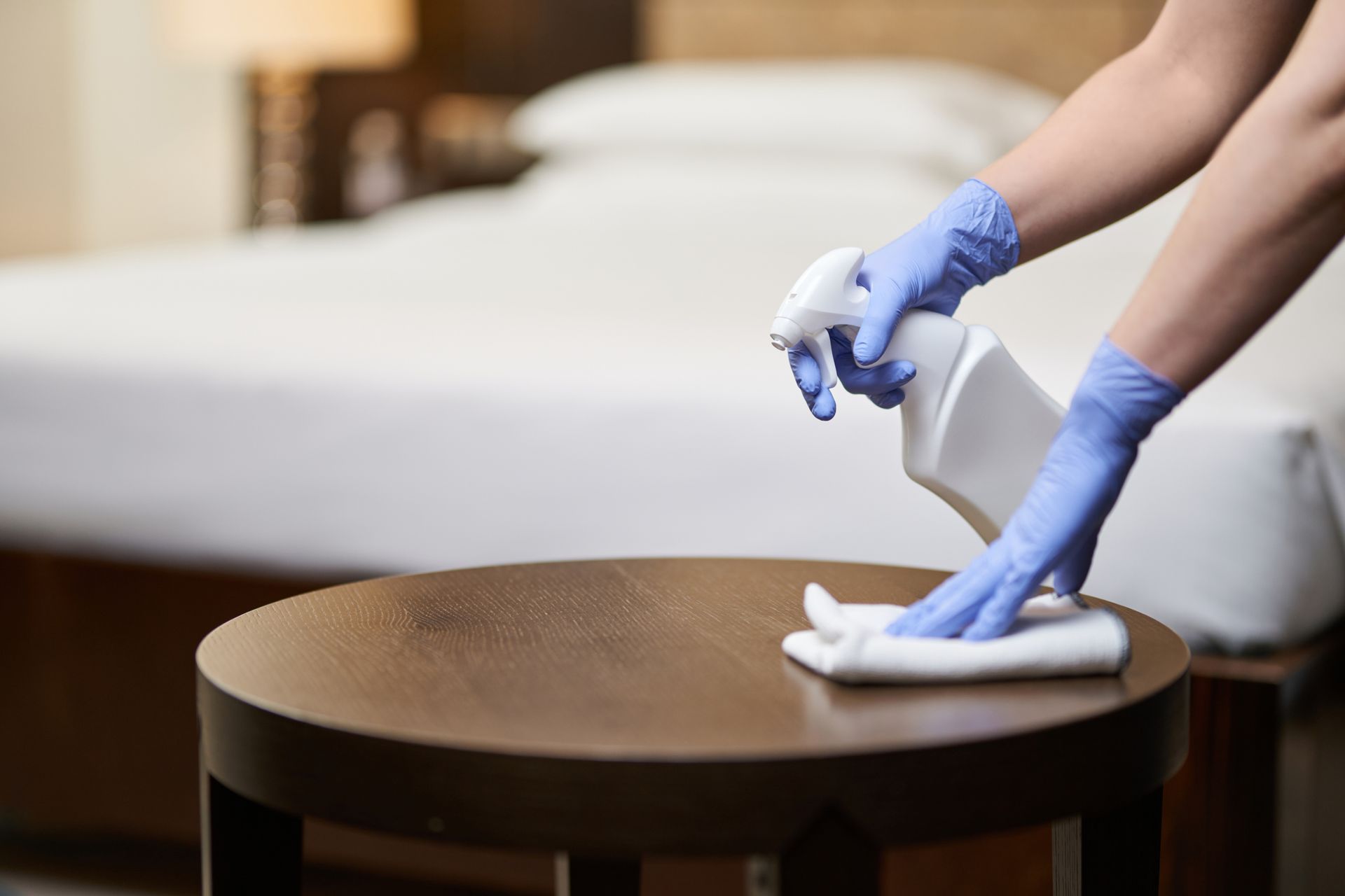 Person in blue gloves sanitizing a dark round table in a hotel room with a white spray bottle and cloth.